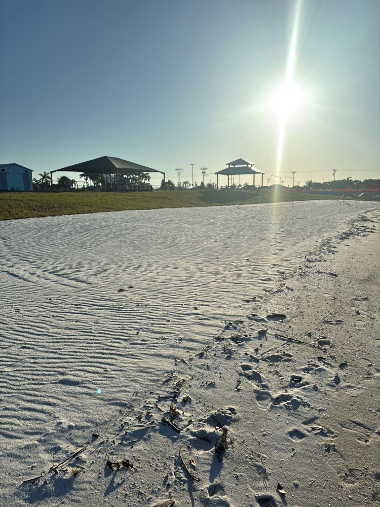 Tropicana Park's sandy beach area, with a large shade structure, a two-tiered gazebo and a small blue building past the grass in the background.